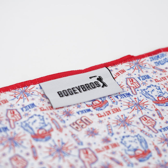 A close-up of the 'Merica - Towel, a microfiber towel with a white BOGEYBROS label and golf swing logo. Its 2-sided print shows USA-themed graphics—fireworks, hands, patriotic phrases—and features a stylish red edge.